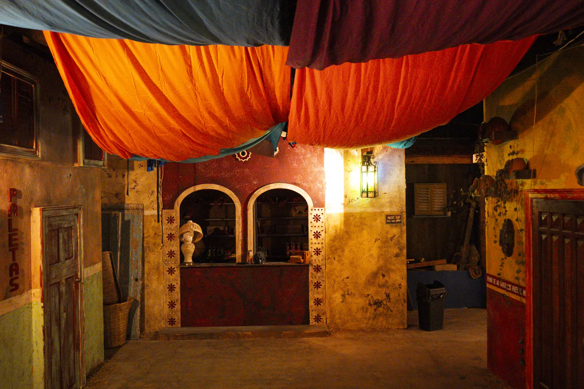 Orange and red fabric canopies over a painted market street, an arched stall counter and a hand-lettered paletas sign