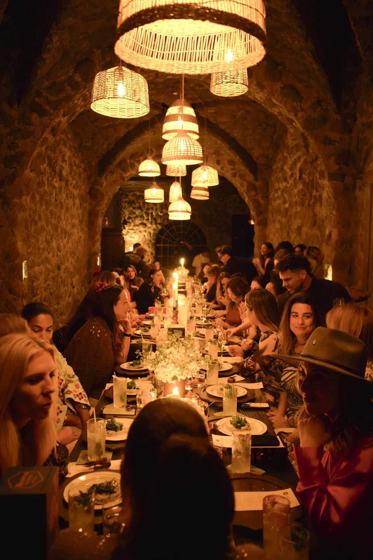Guests at a long candlelit dinner table under woven lamps in the stone cellar of La Cava